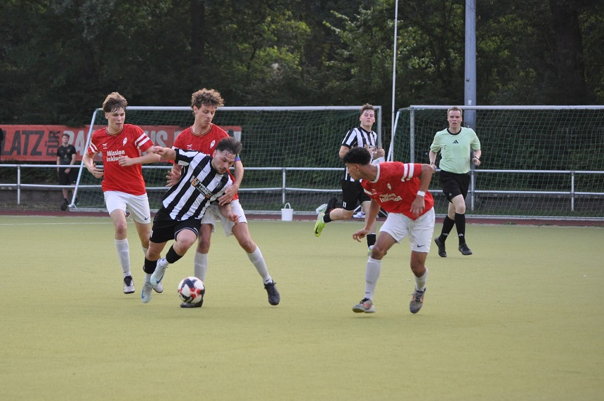 Das Foto zeigt eine Szene eines Fußballspiels auf einem Kunstrasenplatz. Im Vordergrund befinden sich vier Spieler: Drei tragen rote Trikots, einer trägt ein schwarz-weiß gestreiftes Trikot. Der Spieler im gestreiften Trikot führt den Ball am Fuß und wird von zwei Spielern in roten Trikots bedrängt. Ein weiterer Spieler in Rot läuft von links ins Bild.  Im Hintergrund stehen ein Schiedsrichter in einem grünen Trikot und ein Linienrichter, beide tragen schwarze Hosen. Hinter dem Spielfeld ist ein Fußballtor mit Netz zu sehen.