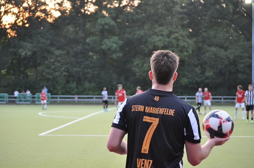 Das Foto zeigt eine Szene eines Fußballspiels auf einem grünen Spielfeld, vermutlich aus der Perspektive eines Zuschauers. Im Vordergrund steht ein Fußballspieler mit dem Rücken zur Kamera. Er trägt ein schwarzes Trikot mit der Aufschrift „STERN MARIENFELDE“, der Rückennummer 7 und dem Namen „VENZ“. In seiner rechten Hand hält er einen Fußball und scheint sich auf einen Einwurf vorzubereiten.
