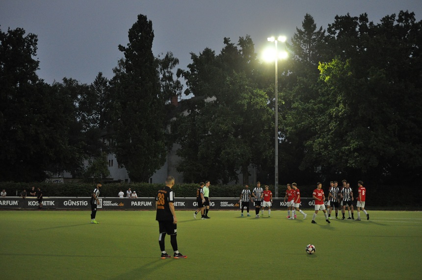 Das Foto zeigt eine Szene eines Fußballspiels auf einem beleuchteten Spielfeld in der Dämmerung oder bei Nacht. Im Vordergrund steht ein Spieler in einem schwarzen Trikot mit der Rückennummer 26. Er scheint sich auf einen Freistoß vorzubereiten. Der Ball liegt vor ihm auf dem Rasen.  Im Hintergrund stehen mehrere Spieler in roten sowie in schwarz-weißen Trikots. Einige von ihnen bilden eine Mauer, wie es bei einem Freistoß üblich ist. Das Spielfeld ist von Bäumen umgeben.