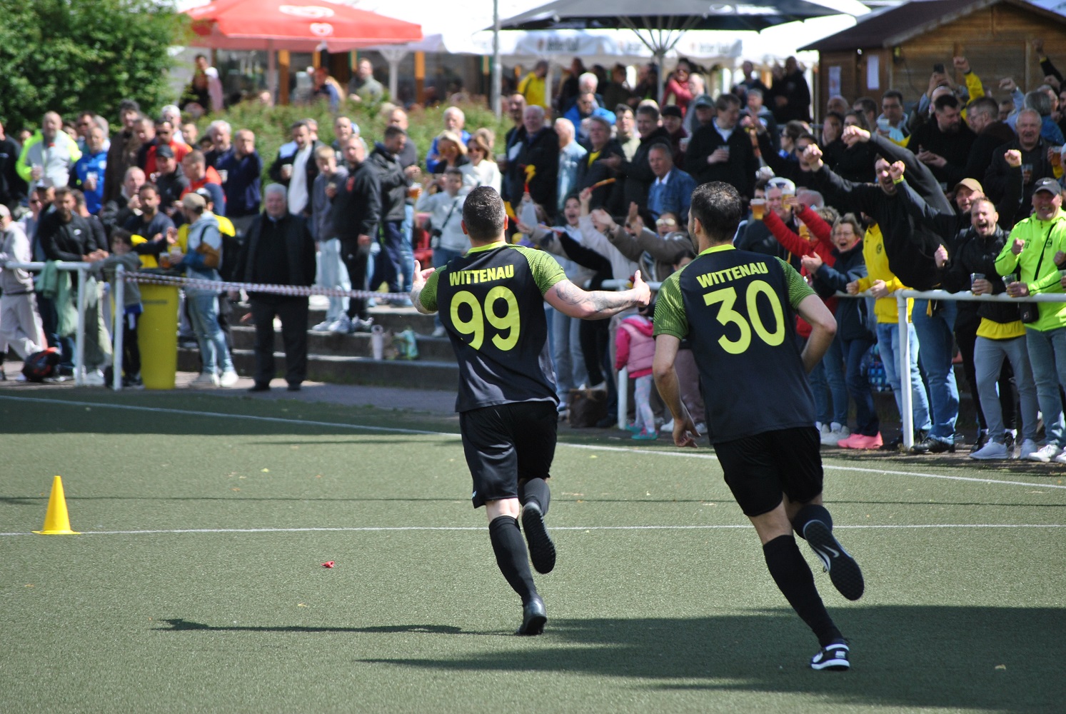 Das Foto zeigt eine Szene auf einem Fußballfeld mit Kunstrasen. Im Vordergrund laufen zwei Sportler*innen mit dem Rücken zur Kamera auf das Spielfeld zu. Beide tragen schwarze Trikots, schwarze Shorts und schwarze Stutzen mit gelben Akzenten. Auf den Trikots stehen in gelber Schrift der Ortsname „Wittenau“ sowie die Rückennummern „99“ und „30“. Die Person mit der Nummer 99 hat die Arme seitlich ausgestreckt, während die Person mit der Nummer 30 leicht nach vorne gebeugt läuft. Am linken Rand des Spielfelds ist ein gelber Trainingskegel zu sehen. Hinter einer niedrigen Absperrung im Hintergrund steht eine große Gruppe von Zuschauer*innen. Viele Menschen tragen Freizeitkleidung, einige halten Getränke in der Hand oder haben die Arme gehoben. Die Gesichter sind teilweise erkennbar, einzelne Personen wirken in Bewegung, andere stehen dicht beieinander. Hinter den Zuschauenden befinden sich Sonnenschirme und kleine Gebäude oder Verkaufsstände, was auf eine Sportveranstaltung im Freien hindeutet. Das Bild vermittelt den Moment eines sportlichen Ereignisses, möglicherweise nach einer Spielsituation oder einem erzielten Tor. Der Fokus liegt auf der Bewegung der beiden Spieler*innen und der dichten Kulisse aus Zuschauenden im Hintergrund.