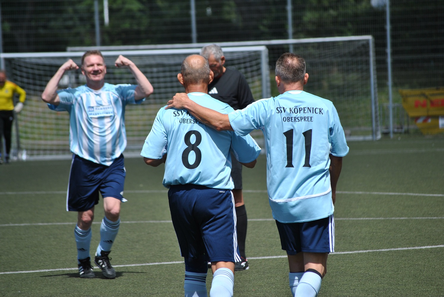 Das Foto zeigt eine Szene auf einem Fußballfeld mit grünem Kunstrasen. Im Vordergrund stehen zwei Sporttreibende mit dem Rücken zur Kamera. Beide tragen hellblaue Trikots, dunkelblaue Shorts und helle Stutzen. Auf den Trikots ist in schwarzer Schrift der Vereinsname „SSV Köpenick Oberspree“ zu lesen, darunter die Rückennummern „8“ und „11“. Die Person mit der Nummer 11 legt einen Arm um die Schultern der Person mit der Nummer 8. Beide bewegen sich gemeinsam über das Spielfeld. Links im Bild läuft eine weitere sporttreibende Person in einem hellblau-weiß gestreiften Trikot und dunklen Shorts auf die beiden zu. Die Person hat beide Arme angewinkelt und hebt sie nach oben. Im mittleren Hintergrund steht eine weitere Person in schwarzer Sportkleidung, teilweise verdeckt. Im Hintergrund ist ein großes Fußballtor mit weißem Rahmen und Netz zu sehen. Dahinter befinden sich Bäume und eine grüne Umgebung, was auf eine Außenanlage hinweist. Die Szene wird von Tageslicht beleuchtet, es sind deutliche Schatten auf dem Boden erkennbar. Das Bild vermittelt einen Moment des gemeinsamen Auftretens auf dem Spielfeld, bei dem mehrere Beteiligte räumlich nah beieinander stehen oder aufeinander zugehen.