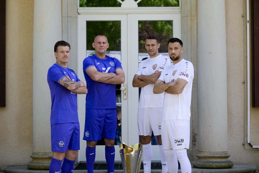 Four people are standing in front of a building with pillars and a glass door. Two of them are wearing blue sportswear, the other two white. All four people are standing with their arms folded around a large cup placed in the middle of the floor. The faces of the people are obscured. The architecture of the building is visible in the background, but no further details or texts can be made out. Photo: Sandra Ritschel