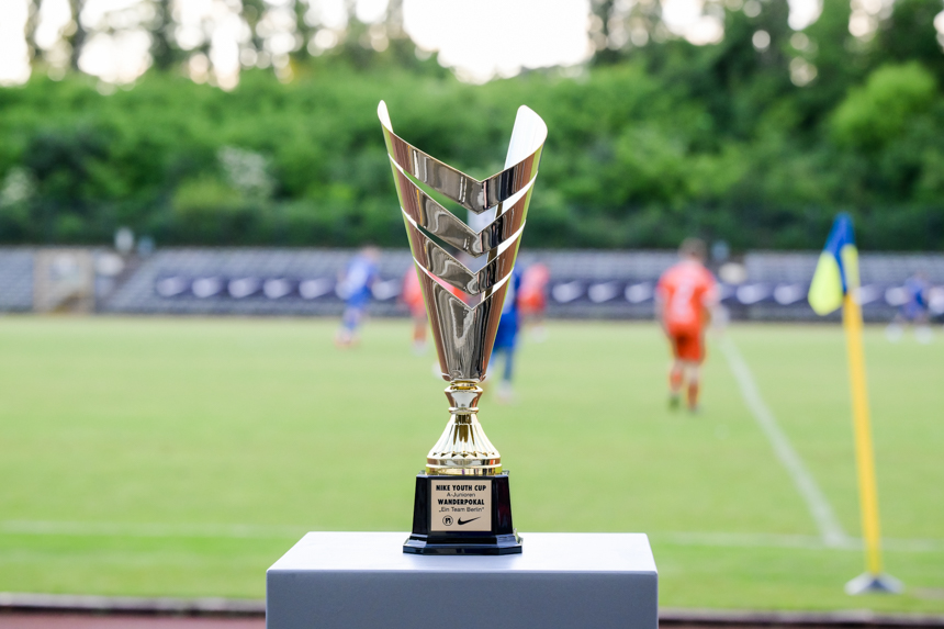 Ein silberner Pokal steht im Fokus auf einem Podest, im Hintergrund ist ein unscharfes Fußballspiel auf grünem Rasen zu sehen. Foto: Sandra Ritschel