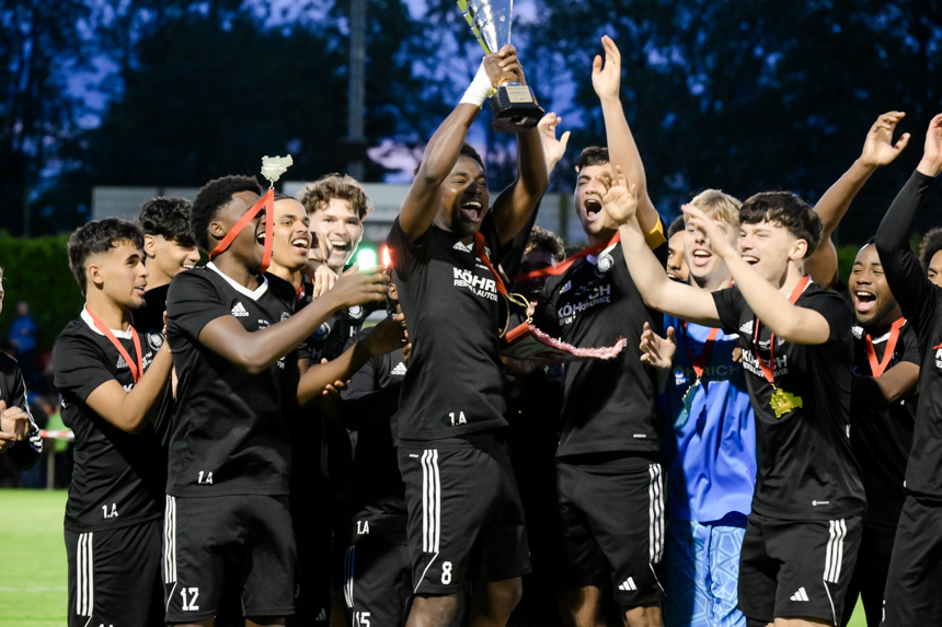 Mehrere Personen in schwarzen Trikots jubeln dicht zusammen, eine Person hält den Pokal hoch, alle tragen Medaillen auf dem Fußballfeld. Foto: Sandra Ritschel