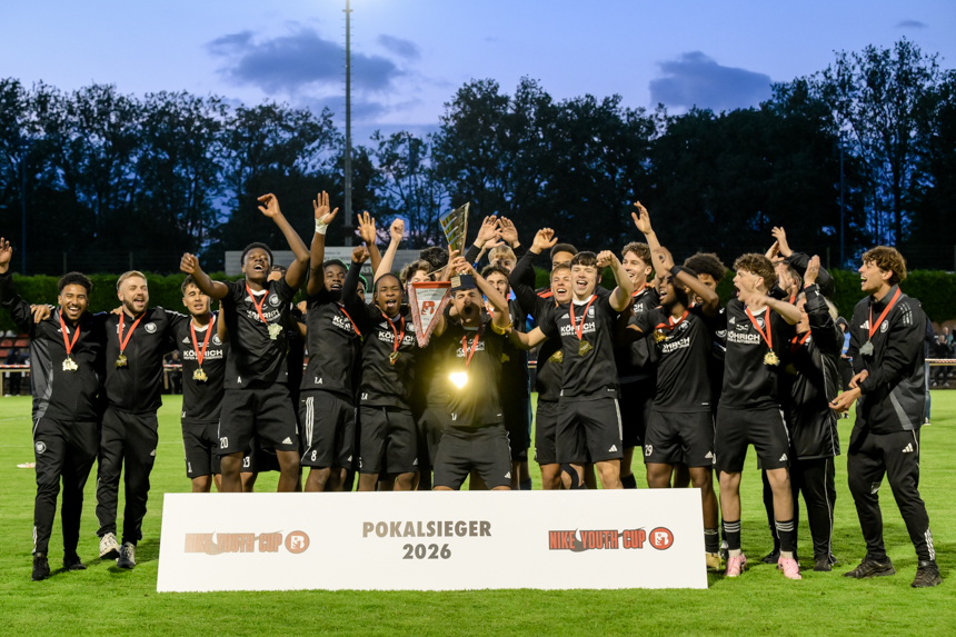 A team in black jerseys cheers with medals and trophy behind a "Cup Winners 2026" banner on the soccer pitch. Photo: Sandra Ritschel