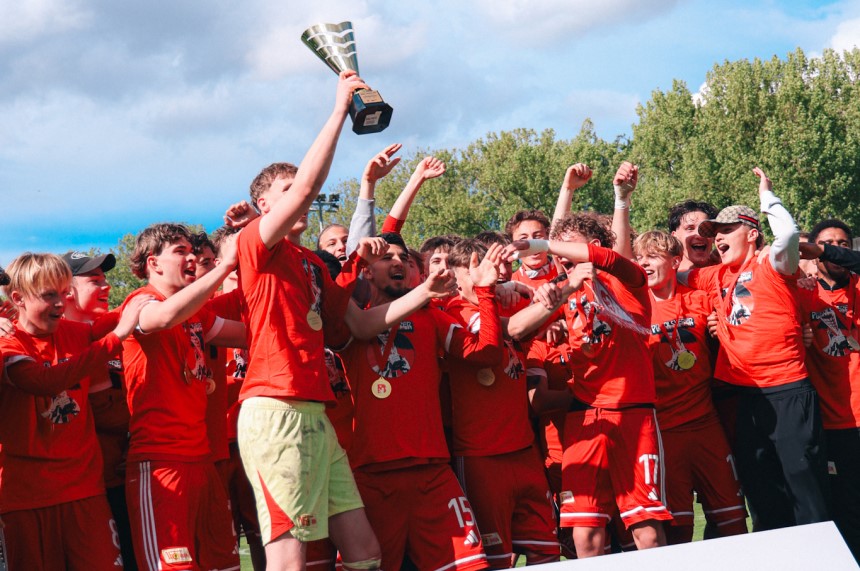 Ein Team in roten Trikots hebt jubelnd einen Pokal, viele tragen Medaillen und stehen dicht zusammen auf einem sonnigen Fußballplatz. Foto: Loredana Zafisambondaoky