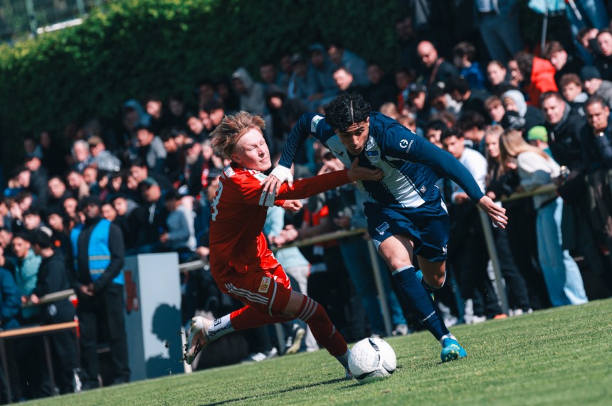 Zwei Spielende in roten und blauen Trikots kämpfen um den Ball, dicht gedrängt vor einer großen Zuschauermenge am Spielfeldrand. Foto: Loredana Zafisambondaoky