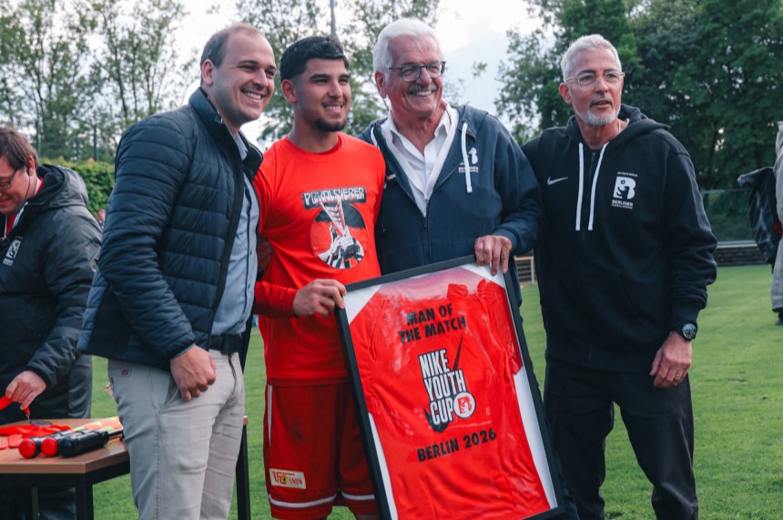 Vier Personen stehen auf dem Rasen und präsentieren ein gerahmtes rotes Trikot mit der Aufschrift „Man of the Match, Berlin 2026“. Foto: Loredana Zafisambondaoky