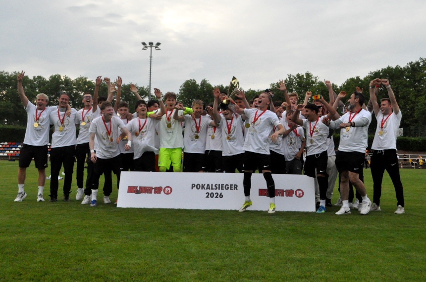 Große Gruppe im Fußballtrikot jubelt auf Rasen, hebt Pokal; Banner „Pokalsieger 2026“, Medaillen sichtbar, gemeinsamer Teamerfolg. Foto: BFV