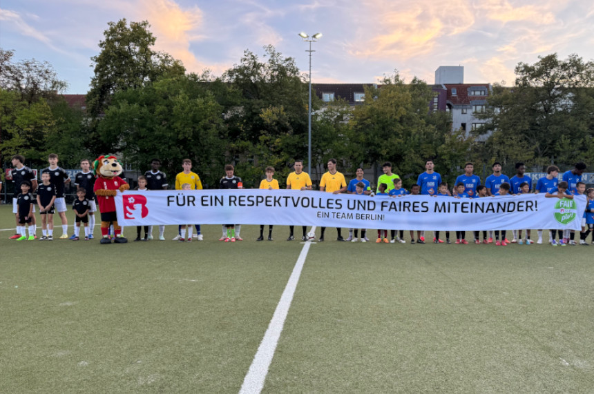 Das Foto zeigt eine Gruppe von etwa 15 Fußballspieler*innen, die gemeinsam auf einem Fußballfeld stehen. Sie halten ein großes, weißes Banner mit der Aufschrift: „FÜR EIN RESPEKTVOLLES UND FAIRES MITEINANDER! EIN TEAM BERLIN!“  Die Spieler*innen tragen unterschiedliche Trikots in verschiedenen Farben. In der Mitte der Gruppe steht eine Person in einem bunten Maskottchenkostüm. Die Gruppe wirkt vielfältig – es sind Menschen unterschiedlichen Alters, Geschlechts und Erscheinungsbildes zu sehen.  Im Hintergrund erkennt man grüne Bäume, einige Gebäude und einen leicht bewölkten Himmel mit einem schönen Sonnenuntergang.