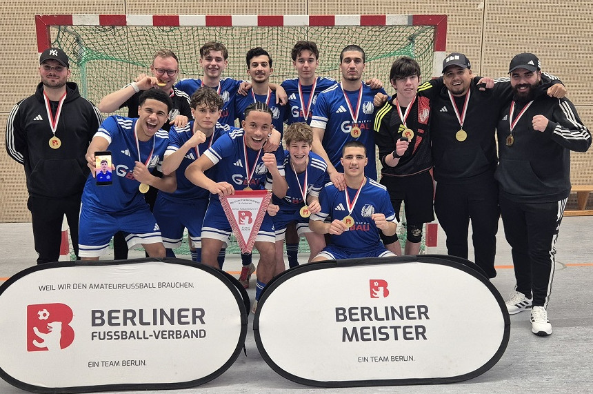 Eine Fußballmannschaft posiert jubelnd in einer Sporthalle vor einem Hallenfußballtor. Die Spieler tragen blaue Trikots, blaue Hosen und blaue Stutzen; an den Seitenlinien stehen mehrere Personen im schwarzen Trainingsanzug, vermutlich Trainer oder Betreuer. Viele in der Gruppe halten Medaillen hoch, zeigen Daumen nach oben oder Siegergesten wie geballte Fäuste. In der Mitte der Vorderreihe präsentiert ein Spieler ein Wimpelbanner des Berliner Fußball-Verbands. Vor der Gruppe stehen zwei ovale Werbetafeln. Die linke trägt den Text: „Weil wir den Amateurfußball brauchen… Berliner Fußball-Verband – Ein Team Berlin“. Die rechte zeigt: „Berliner Meister – Ein Team Berlin“, jeweils mit rotem Verbandslogo. Die Stimmung wirkt ausgelassen und stolz, als feiere das Team einen frisch gewonnenen Meistertitel oder Turniererfolg. Die Halle hat helle Wände und einen Boden aus Sportbelag, typisch für Indoor-Fußballspielflächen.