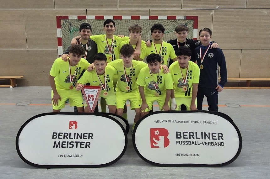 Eine Fußballmannschaft steht in einer Sporthalle dicht beisammen vor einem Hallenfußballtor. Die Spieler tragen leuchtend gelbe Trikots und Hosen, während eine Person am rechten Rand ein dunkelblaues Trainingsoutfit trägt – vermutlich ein Trainer oder Betreuer. Viele halten Medaillen in der Hand oder zeigen sie stolz nach vorne. In der vorderen Reihe hält ein Spieler einen Wimpel des Berliner Fußball-Verbands. Vor dem Team stehen zwei ovale Werbetafeln. Links steht „Berliner Meister – Ein Team Berlin“, rechts „Berliner Fußball-Verband – Ein Team Berlin“, jeweils mit rotem Verbandslogo. Die Gruppe wirkt freudig und stolz, als hätte sie einen Meistertitel oder eine große sportliche Leistung erreicht. Die Umgebung ist eine typische Sporthalle mit hellem Wandbelag und orangenen Markierungen auf dem Hallenboden.