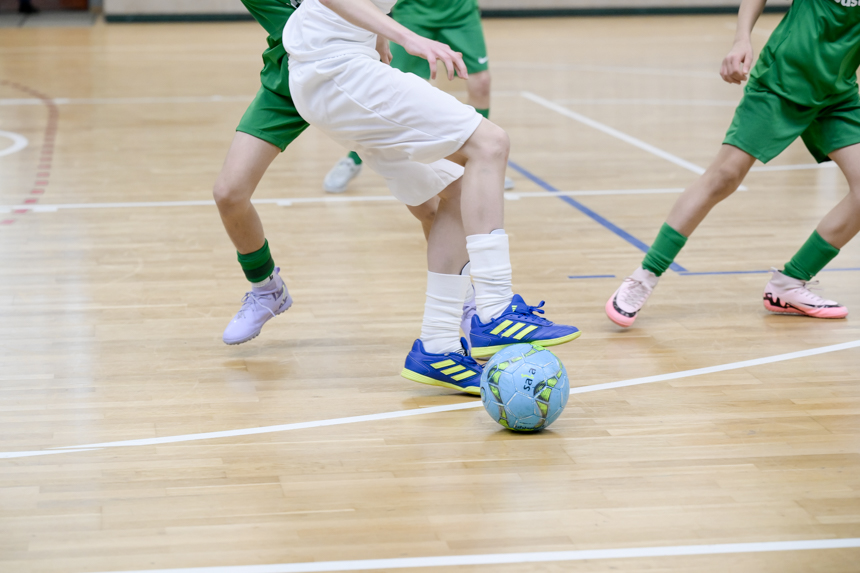 The picture shows a scene in a sports hall with a light-colored wooden floor and white playing field lines. Three people can be seen actively playing soccer in the foreground. The people are only partially visible - mainly their legs and feet. One person is wearing white shorts, white socks and blue sports shoes with yellow stripes. This person is standing right next to the ball, which is light blue and has green and black patterns. Two other people are wearing green jerseys and green shorts. They move dynamically around the ball as if they are trying to capture it. Their shoes are different: one pair is pink, the other light purple. The background is blurred, but you can make out the boundary lines of the pitch and part of the hall wall.