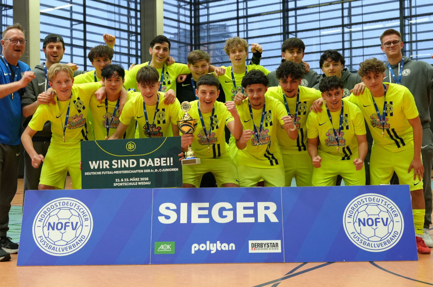 Das Foto zeigt eine große Gruppe von jungen Sportlerinnen und Sportlern, die gemeinsam in einer Sporthalle posieren. Die Gruppe steht dicht beieinander und bildet zwei Reihen. Alle Personen tragen einheitliche, leuchtend gelbe Sportkleidung mit dunklen Streifen und haben Medaillen um den Hals hängen. Vor der Gruppe steht ein breites, blaues Schild mit der Aufschrift „SIEGER“. Auf dem Schild befinden sich außerdem mehrere Logos, darunter das Logo des Nordostdeutschen Fußballverbands (NOFV) sowie Sponsorenlogos. In der Mitte der vorderen Reihe hält eine Person eine Trophäe in der Hand. Direkt daneben wird ein großes, grünes Schild hochgehalten. Links und rechts neben der Gruppe stehen zwei Menschen in blauer oder grauer Sportkleidung, die ebenfalls mitjubeln. Der Hintergrund zeigt eine helle Sporthalle mit hohen Fenstern und dunklen Wandbereichen. Der Boden besteht aus typischem Hallenbelag mit Linien für verschiedene Sportarten. Die gesamte Gruppe zeigt eine feierliche Siegerpose, teilweise mit erhobenen Armen oder geballten Fäusten.