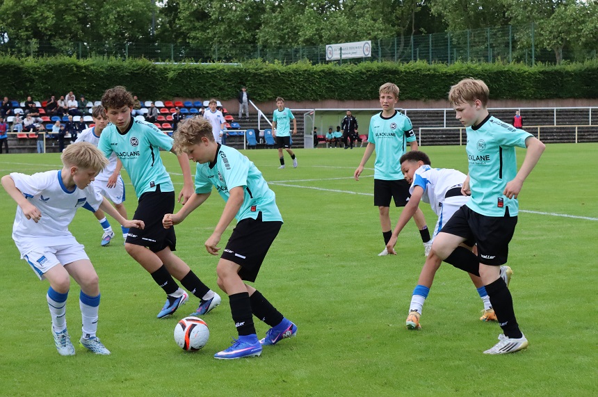 Das Foto zeigt eine lebhafte Spielsituation auf einem Fußballfeld im Freien. Der Rasen ist gleichmäßig grün und wirkt gepflegt. Mehrere junge Personen sind in der Mitte des Bildes eng beieinander und konzentrieren sich auf den Fußball, der sich am Boden im Zentrum der Szene befindet. Im Vordergrund, leicht links der Bildmitte, stehen zwei Personen aus unterschiedlichen Teams dicht nebeneinander und versuchen, den Ball zu kontrollieren oder zu erobern. Eine Person trägt ein weißes Trikot mit blauen Details, weiße Shorts und helle Fußballschuhe. Direkt daneben befindet sich eine Person mit helltürkisem Trikot, schwarzen Shorts und dunklen Socken, die mit einem ausgestreckten Bein ebenfalls auf den Ball einwirkt. Der Ball ist ein moderner Fußball mit weißen Flächen und auffälligen schwarzen sowie orangefarbenen Elementen. Um diese zentrale Aktion herum gruppieren sich mehrere weitere Personen in ähnlichen helltürkisen Trikots sowie einzelne in weißen Trikots. Eine Person im rechten Vordergrund hebt ein Bein an, als würde ein Pass vorbereitet oder der Ball abgefangen werden. Eine andere Person im rechten mittleren Bereich ist leicht nach vorne gebeugt und nah am Boden, was darauf hindeutet, dass diese Person gerade ins Straucheln geraten ist oder versucht, das Gleichgewicht zu halten. Im Mittelfeldbereich des Bildes stehen weitere Mitspielende mit etwas Abstand zur direkten Spielsituation und beobachten oder positionieren sich für den nächsten Spielzug. Alle tragen typische Fußballbekleidung wie Trikots, Shorts, lange Socken und Fußballschuhe. Im Hintergrund ist eine kleine Tribüne mit mehrfarbigen Sitzschalen (rot, blau und weiß) zu sehen, auf der einige Zuschauende sitzen. Dahinter verläuft eine Hecke und darüber hinaus sind Bäume sichtbar, die den Platz einfassen. Seitlich des Spielfelds erkennt man außerdem eine Trainerbank oder Auswechselbereich mit weiteren Personen. Das Licht wirkt natürlich, vermutlich Tageslicht bei leicht bewölktem Himmel, wodurch die Farben klar, aber nicht übermäßig kontrastreich erscheinen. Die Szene insgesamt vermittelt Dynamik durch die enge Gruppierung um den Ball und die verschiedenen Bewegungsrichtungen der beteiligten Personen.