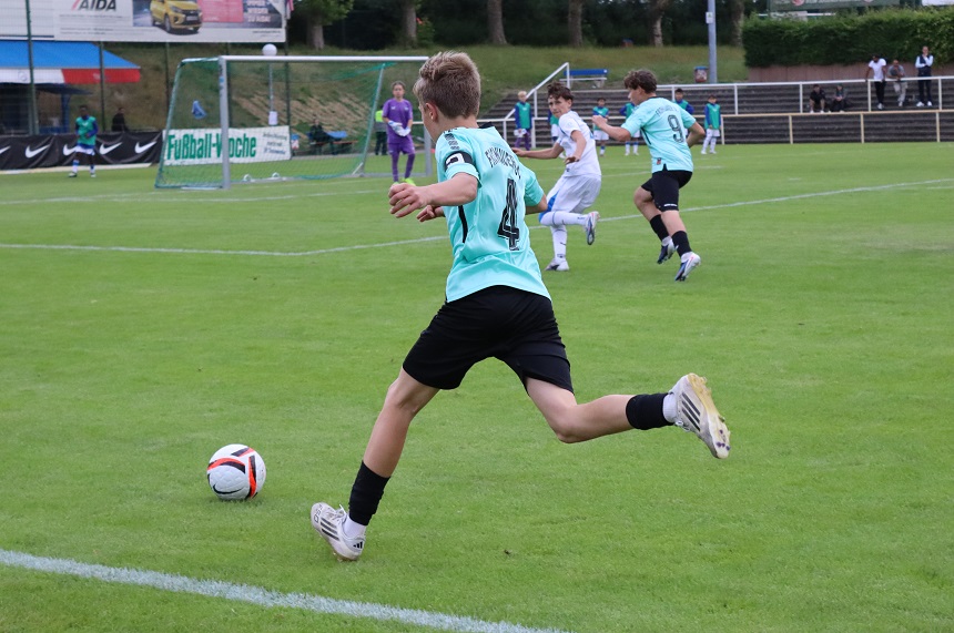 Das Foto zeigt eine Szene aus einem Fußballspiel auf einem grünen Rasenplatz im Freien. Im Vordergrund befindet sich eine Person in einem helltürkisen Trikot mit der schwarzen Rückennummer „4“, schwarzen Shorts, schwarzen Socken und hellen Fußballschuhen. Diese Person läuft mit ausgestrecktem rechten Bein auf einen Fußball zu, der sich knapp vor der rechten Fußspitze befindet. Die Körperhaltung deutet darauf hin, dass gleich ein Pass oder Schuss ausgeführt wird. Im mittleren Bildbereich sind mehrere weitere Personen zu sehen, die ebenfalls am Spiel beteiligt sind. Zwei Personen tragen ähnliche helltürkise Trikots und bewegen sich in Richtung Spielfeldmitte. Eine weitere Person in einem weißen Trikot läuft in die gleiche Richtung. Alle tragen typische Fußballausrüstung. Im Hintergrund befindet sich ein Fußballtor mit Netz. Davor steht eine Person in einem dunkleren Outfit, vermutlich auf der Torwartposition. Hinter dem Tor sind Werbebanden mit teilweise lesbarem Text angebracht, unter anderem „Fußball…“ und ein Logo sowie ein Plakat mit dem Schriftzug „AIDA“. Weiter hinten sieht man eine niedrige Tribüne oder Sitzstufen mit einigen zuschauenden Personen sowie Bäume und Büsche, die das Spielfeld einrahmen. Die Szene ist bei Tageslicht aufgenommen. Die Farben sind natürlich, mit einem starken Kontrast zwischen dem satten Grün des Rasens und den hellen sowie dunklen Farben der Trikots. Die Aufnahme wirkt dynamisch, da mehrere Bewegungsrichtungen gleichzeitig sichtbar sind und der Ball im Fokus des Spiels steht.