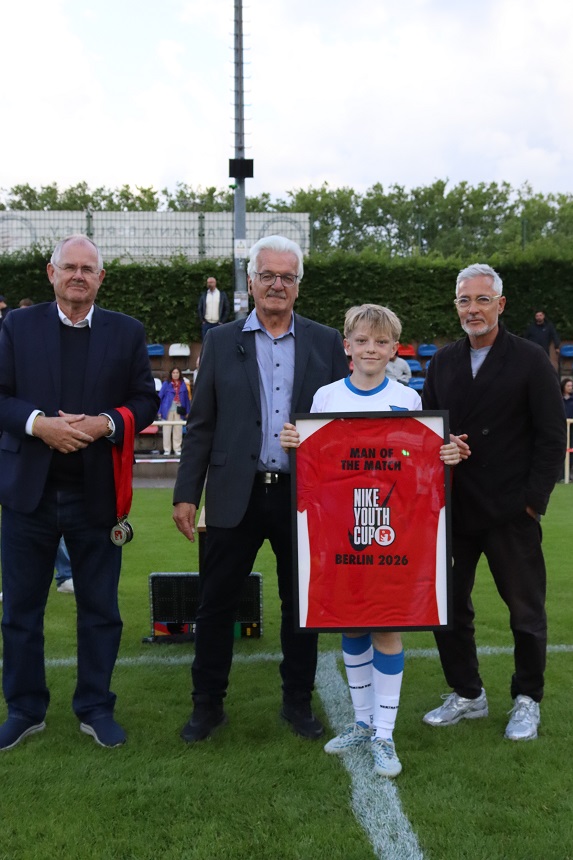 Das Foto zeigt eine Szene auf einem Fußballplatz während einer Auszeichnung oder Ehrung. Im Vordergrund stehen vier Personen nebeneinander auf dem Rasen. Die Gruppe ist mittig im Bild positioniert, mit einem Fußballfeld und Zuschauerbereich im Hintergrund. Die zweite Person von rechts hält ein gerahmtes rotes Trikot vor dem Körper. Das Trikot ist deutlich sichtbar und enthält schwarzen Text. Auf dem Trikot steht: „MAN OF THE MATCH NIKE YOUTH CUP BERLIN 2026“ Das Trikot ist in einen schwarzen Rahmen eingefasst und wirkt wie ein Präsentations- oder Erinnerungsstück. Die Person, die das Trikot hält, trägt ein sportliches Outfit mit weißem Trikot, weißen Shorts, weißen Socken und Fußballschuhen. Die drei anderen Personen tragen Alltags- beziehungsweise formellere Kleidung: dunkle Jacken oder Sakkos, lange Hosen und geschlossene Schuhe. Eine Person ganz links hält zusätzlich einen Gegenstand mit einem Band in den Händen, der an eine Medaille oder Auszeichnung erinnert. Alle vier Personen stehen dicht nebeneinander und sind zur Kamera ausgerichtet. Die Füße befinden sich auf einer weißen Linie des Spielfelds, was darauf hindeutet, dass die Aufnahme direkt auf dem Platz gemacht wurde. Im Hintergrund sind weitere Elemente eines Sportgeländes sichtbar: eine niedrige Tribüne mit bunten Sitzplätzen, einzelne Zuschauerinnen und Zuschauer, eine Hecke sowie Bäume. Außerdem steht hinter der Gruppe ein hoher Mast, vermutlich für Flutlicht oder Technik. Die Aufnahme ist bei Tageslicht entstanden. Die Farben wirken natürlich, mit einem deutlichen Kontrast zwischen dem grünen Rasen, dem roten Trikot im Rahmen und den dunkleren Farbtönen der Kleidung. Die Szene vermittelt den Eindruck einer offiziellen Ehrungsmomentaufnahme im Anschluss oder im Rahmen eines Fußballspiels.