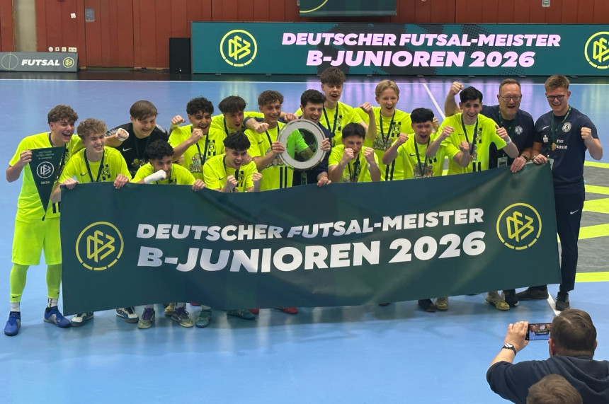 Das Foto zeigt eine Gruppe von Personen auf einem Hallenspielfeld, die eng zusammenstehen und gemeinsam posieren. Alle tragen leuchtend gelbe Sportkleidung, während zwei Personen am rechten Bildrand dunkle Trainingskleidung tragen. Die Gruppe hält ein großes, dunkelgrünes Banner vor sich. Auf dem Banner steht in weißen und gelben Buchstaben: „DEUTSCHER FUTSAL-MEISTER B-JUNIOREN 2026“ Links und rechts auf dem Banner befindet sich das Logo des Deutschen Fußball-Bundes (DFB). In der Mitte der Gruppe hält eine Person einen runden Pokal oder eine Trophäe in die Höhe. Mehrere Personen tragen Medaillen um den Hals. Der Hallenboden ist hellblau, und im Hintergrund hängt ein weiteres großes Banner mit der gleichen Aufschrift wie auf dem Frontbanner. Auf der linken Seite erkennt man zusätzlich ein kleines Logo des Futsal-Wettbewerbs. Im Vordergrund rechts kniet eine Person, die mit einem Handy oder einer Kamera ein Foto oder Video der Gruppe aufnimmt. Die Szene vermittelt den Moment einer Siegerehrung nach einem sportlichen Wettbewerb.