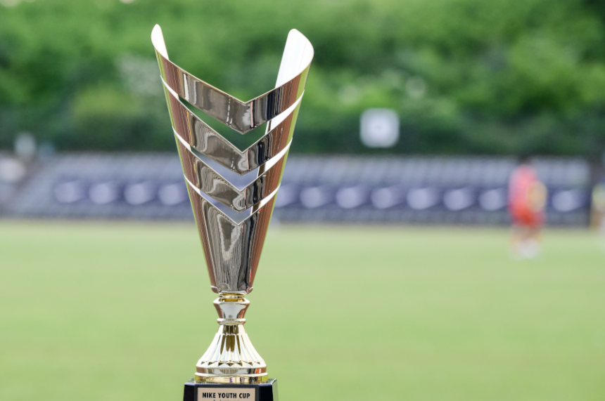 The focal point of the photo is a tall, silver-colored trophy made of shiny metal. The trophy tapers towards the bottom and is made of several V-shaped elements arranged on top of each other, which are open at the top. It stands on a small black base with a gold-colored plaque. The text "NIKE YOUTH CUP" can be read on the plaque. The background is blurred and shows a green lawn reminiscent of a sports field. Behind it, dark rows of seats can be seen, like in a stadium. A blurred, standing person in sportswear can be seen on the right edge of the picture. Due to the shallow depth of field, the visual focus is clearly on the trophy.