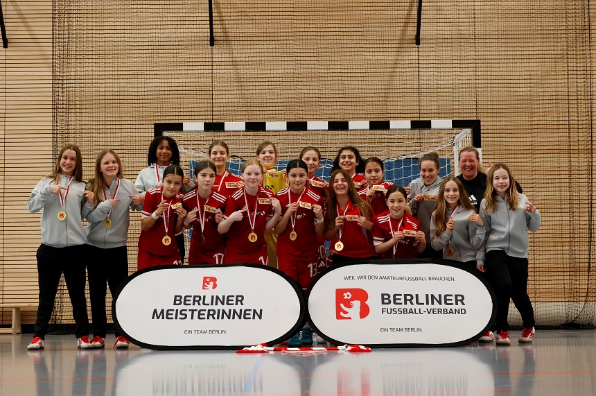 Eine Mädchen- oder Frauenfußballmannschaft steht dicht beisammen in einer Sporthalle vor einem Hallenfußballtor. Die Spielerinnen tragen rote Trikots, rote Hosen und rote Stutzen. Einige Personen am linken und rechten Rand tragen graue Kapuzenpullover und wirken wie Trainerinnen oder Betreuerinnen. In der Mitte hält eine Spielerin ein Wimpelbanner des Berliner Fußball-Verbands hoch. Viele Teammitglieder tragen Goldmedaillen um den Hals und zeigen sie stolz nach vorne. Die Stimmung ist fröhlich und ausgelassen, als feiere die Gruppe einen frisch gewonnenen Meistertitel. Im Vordergrund stehen zwei große ovale Werbetafeln. Die linke trägt den Schriftzug: „Berliner Meisterinnen – Ein Team Berlin.“ Die rechte zeigt: „Berliner Fußball-Verband – Ein Team Berlin.“ Beide Tafeln sind weiß mit rotem Logo. Die Sporthalle hat helle, holzfarbene Wände und einen glänzenden Hallenboden. Das grün-weiß markierte Tor im Hintergrund deutet auf ein Indoor-Fußballturnier hin.