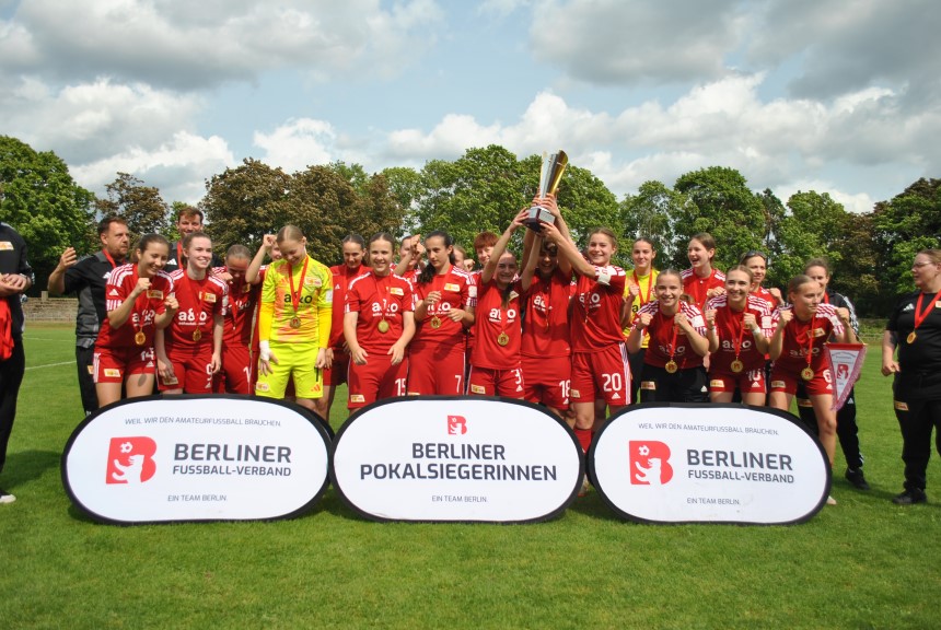 Eine Fußballmannschaft in roten Trikots feiert mit Pokal. Zwei Banner zeigen „Berliner Pokalsiegerinnen“ und „Berliner Fußball-Verband“. Foto: BFV