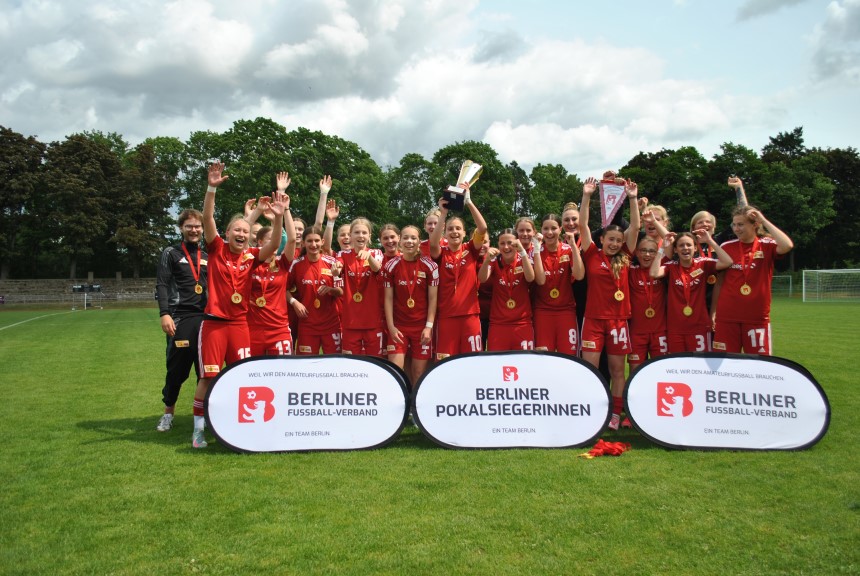 Eine Fußballmannschaft in roten Trikots feiert mit Pokal. Zwei Banner zeigen „Berliner Pokalsiegerinnen“ und „Berliner Fußball-Verband“. Foto: BFV