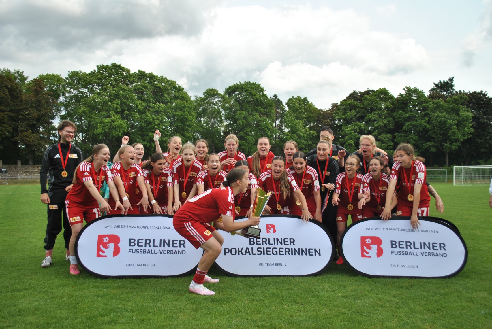 Eine Fußballmannschaft in roten Trikots feiert mit Pokal. Zwei Banner zeigen „Berliner Pokalsiegerinnen“ und „Berliner Fußball-Verband“. Foto: BFV