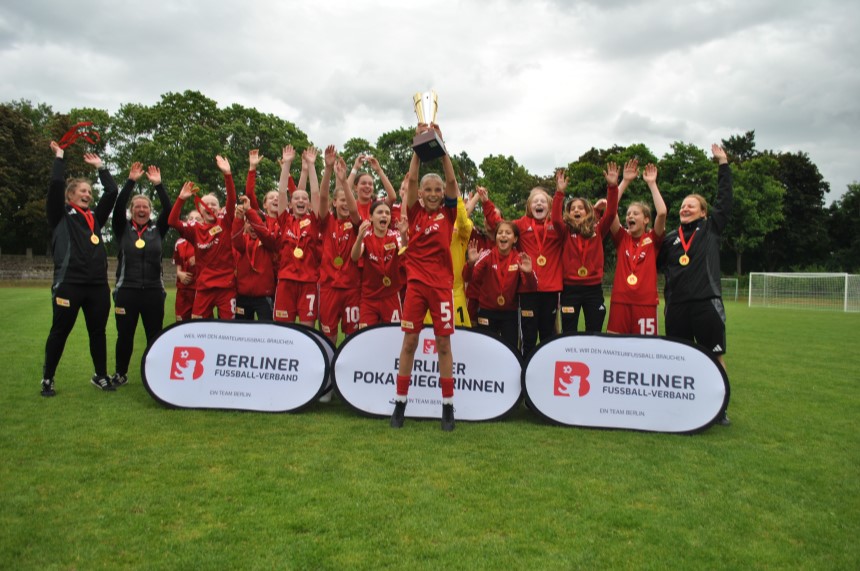 Eine Fußballmannschaft in roten Trikots feiert mit Pokal. Zwei Banner zeigen „Berliner Pokalsiegerinnen“ und „Berliner Fußball-Verband“. Foto: BFV