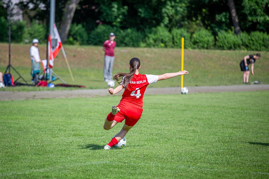 Auf dem Bild ist ein Fußballspiel zu sehen. Im Vordergrund tritt eine Person im roten Trikot mit der Nummer 4 und der Aufschrift „TEAM BERLIN“ einen Ball. Im Hintergrund beobachten mehrere Menschen das Spiel oder stehen am Spielfeldrand. Foto: Joseph Narbey