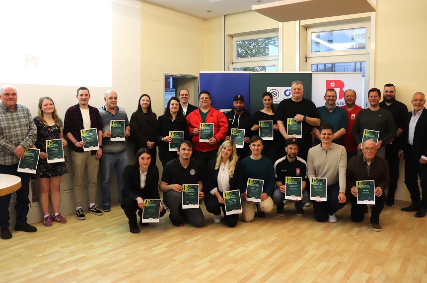 Das Foto zeigt eine Gruppe von etwa 30 erwachsenen Personen, die in zwei Reihen in einem hellen Innenraum stehen und knien. Der Raum wirkt wie ein Veranstaltungs‑ oder Seminarraum mit hellem Holzboden, weißen Wänden und großen Fenstern im Hintergrund. Tageslicht fällt von der rechten Seite in den Raum. Im Mittelpunkt stehen die Personen, die fast alle eine gerahmte Urkunde oder ein Zertifikat mit dunklem Hintergrund und grünen Elementen in den Händen halten. Die Tafeln sind überwiegend im Hochformat. Die Personen in der vorderen Reihe knien oder hocken, während die Personen in der hinteren Reihe stehen. Alle blicken in Richtung Kamera. Die Kleidung ist unterschiedlich: Sie reicht von sportlich über casual bis hin zu schlichter Business‑Kleidung. Zu sehen sind verschiedene Oberteile wie Pullover, Kapuzenjacken, Hemden, T‑Shirts und Jacken in Farben wie Schwarz, Grau, Blau, Grün, Rot und Beige. Im Hintergrund stehen mehrere Roll‑ups oder Banner mit Logos und Symbolen in Blau, Grün, Weiß und Rot. Der Text auf den Bannern ist teilweise sichtbar, aber nicht vollständig lesbar. Die Banner deuten auf eine organisierte Veranstaltung oder eine Ehrung hin. Der Gesamteindruck des Bildes ist eine formelle Gruppenaufnahme im Rahmen einer Auszeichnung oder Anerkennung, bei der viele Teilnehmende gemeinsam präsentiert werden.