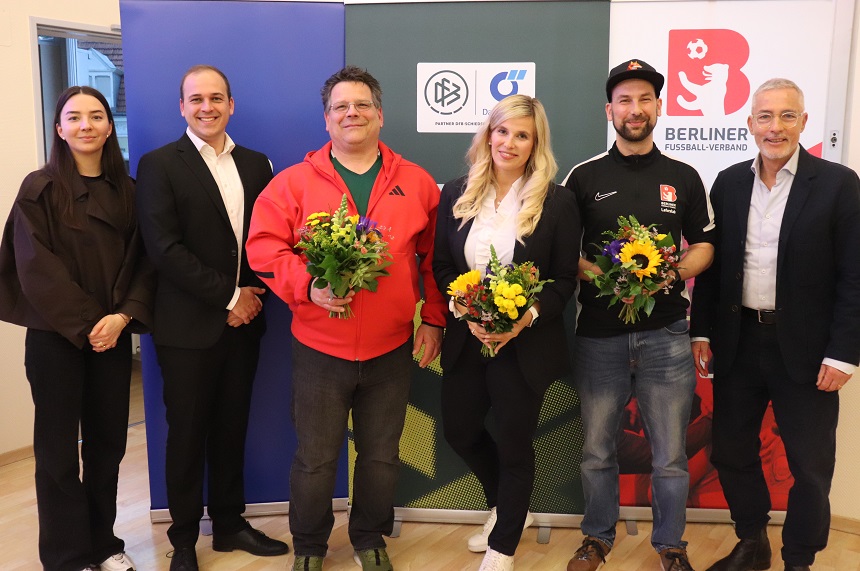 Das Foto zeigt sieben erwachsene Personen, die nebeneinander in einer Reihe in einem hellen Innenraum stehen. Der Boden ist aus hellem Holz, die Wände sind neutral gehalten. Die Personen stehen vor mehreren aufgestellten Roll‑ups oder Bannern. Drei der Personen halten Blumensträuße in den Händen. Die Blumen bestehen aus gelben, grünen und orangefarbenen Blüten mit grünen Blättern. Die übrigen Personen stehen mit den Armen locker am Körper oder vor dem Körper verschränkt. Die Kleidung ist unterschiedlich und reicht von formeller Business‑Kleidung wie Anzügen und Blazern bis zu sportlicher oder legerer Kleidung wie Pullover, Kapuzenjacke, T‑Shirt und Jeans. Die Farben der Kleidung sind unter anderem Schwarz, Dunkelblau, Rot, Weiß und Grau. Im Hintergrund sind mehrere Banner mit Logos und Texten zu sehen. Auf einem Banner ist deutlich der Schriftzug „Berliner Fußball‑Verband“ sowie ein rotes Logo mit einem stilisierten Tier zu erkennen. Auf weiteren Bannern sind grafische Symbole und Abkürzungen sichtbar, der vollständige Text ist jedoch nicht komplett lesbar. Alle Personen schauen in Richtung Kamera. Die Aufnahme wirkt wie ein offizielles Gruppenfoto im Rahmen einer Veranstaltung oder Ehrung in einem sportlichen oder verbandlichen Kontext.