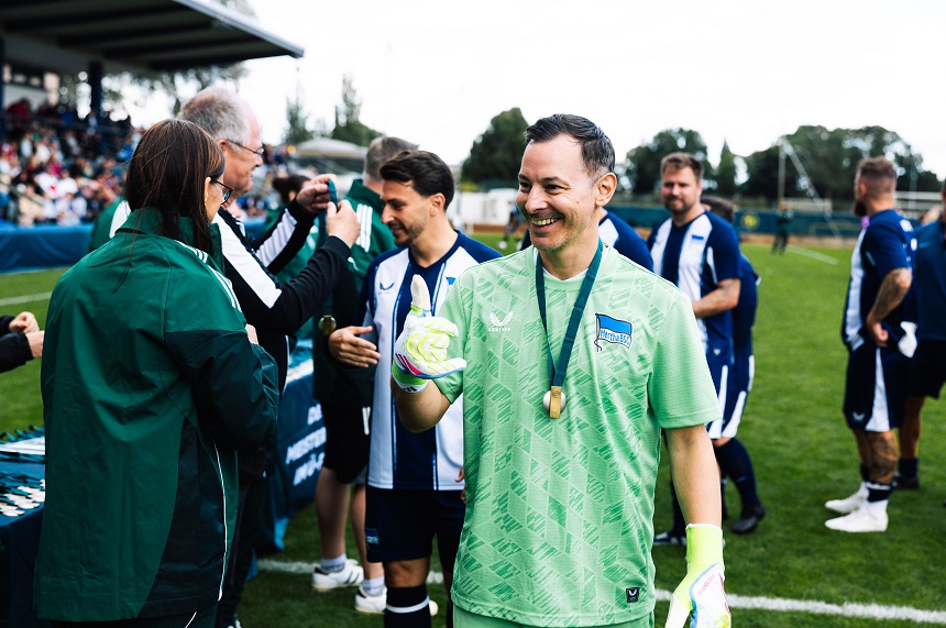 Auf einem Fußballplatz findet eine Siegerehrung statt. Im Vordergrund steht eine Person in einem hellgrünen Torwarttrikot mit passenden Torwarthandschuhen und einer Medaille um den Hals. Die Person zeigt eine Handgeste mit erhobenem Daumen. Im Hintergrund stehen weitere Personen in blau-weißer Sportkleidung sowie Personen in grünen Jacken an einem Ausgabetisch, an dem Medaillen überreicht werden. Links ist eine Tribüne mit zahlreichen Zuschauenden zu sehen, rechts erstreckt sich die Rasenfläche des Spielfelds. Die Szene spielt bei Tageslicht und vermittelt einen Moment der Anerkennung im Rahmen eines Fußballturniers. Die Farben Grün, Blau und Weiß prägen das Bild.