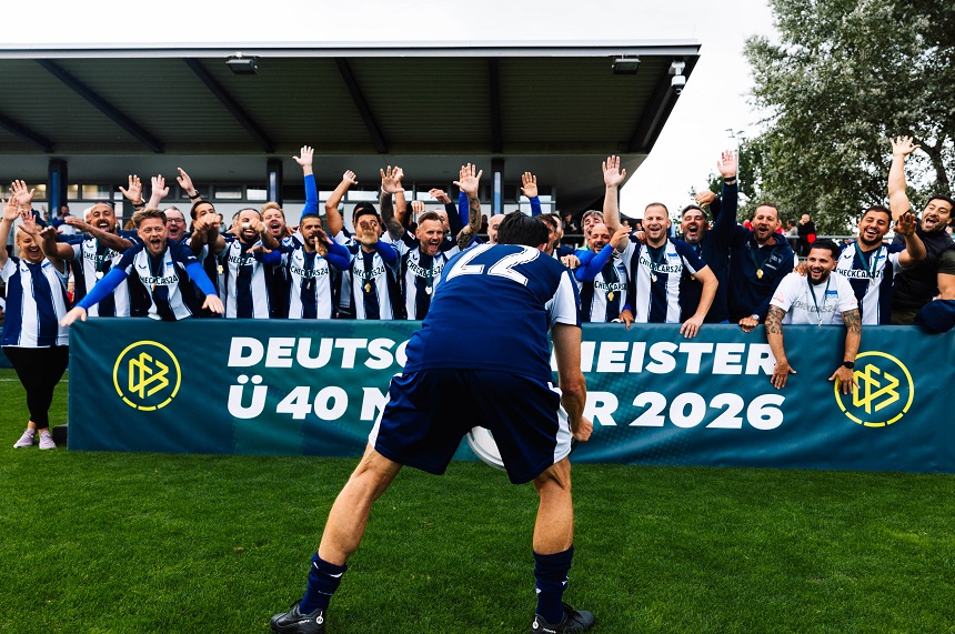 Eine größere Gruppe von Personen in blau-weiß gestreiften Fußballtrikots steht auf einem Fußballplatz hinter einer Barriere und hebt die Arme. Im Vordergrund steht eine Person mit dem Rücken zur Kamera in dunkelblauer Sportkleidung mit der Rückennummer 22. Zwischen der Person im Vordergrund und der Gruppe befindet sich eine grüne Werbebande mit dem DFB-Logo und der gut lesbaren Aufschrift: „Deutscher Meister Ü 40 Männer 2026“. Das Foto wurde auf einer Rasenfläche vor einer überdachten Tribüne aufgenommen. Im Hintergrund sind weitere Personen, Bäume und Teile der Sportanlage zu sehen. Die Farben Blau, Weiß und Grün prägen das Bild. Die Szene zeigt einen gemeinsamen Moment im Umfeld eines Fußballwettbewerbs.