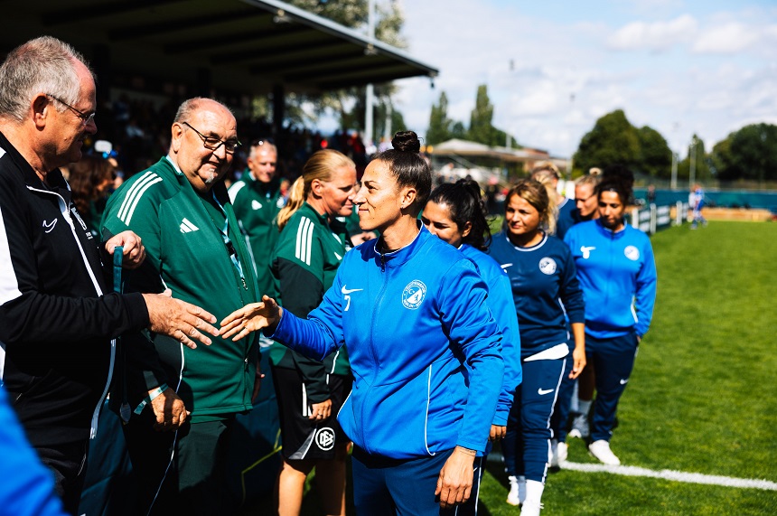 Mehrere Personen in Sportkleidung stehen auf einem Fußballplatz in einer Reihe und begrüßen sich per Handschlag. Im Vordergrund reicht eine Person in einem blauen Trainingsanzug einer Person in dunkler Sportkleidung die Hand. Dahinter warten weitere Personen in blauen und grünen Trainingsanzügen entlang der Seitenlinie des Spielfelds. Im Hintergrund sind eine überdachte Tribüne mit Zuschauenden, Bäume sowie weitere Bereiche der Sportanlage zu sehen. Die Szene spielt bei Tageslicht auf einem grünen Rasenplatz und vermittelt einen fairen und respektvollen Austausch im Rahmen eines Fußballturniers der Altersklasse Ü32. Die Farben Blau, Grün und Schwarz prägen das Bild.