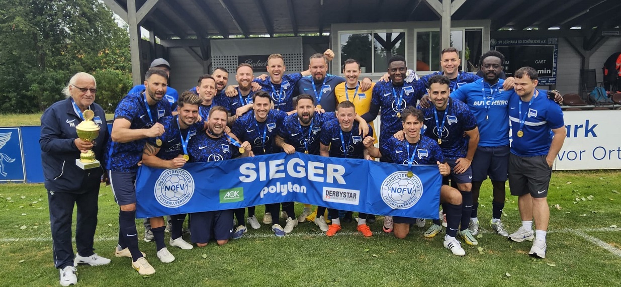 The photo shows a large group of people on a soccer field in front of a club building with a covered seating area. Most are wearing dark blue jerseys and practice gear with a blue patterned top. Many have medals around their necks. The group is arranged in two rows. Some people are standing, while others are kneeling or leaning forward. A blue banner is being held in the foreground. It reads “SIEGER” in large white letters. Also visible are the logos of the Northeast German Soccer Association (NOFV), AOK, Polytan, and Derbystar.  On the left side of the image, a person is standing with a large gold-colored trophy. Additional medals are visible within the group. Most of the people are looking toward the camera.  In the background is a clubhouse or grandstand building with wooden supports and windows. A scoreboard can be seen on the back wall. Advertising banners run along the edge of the field on the right and left. Trees and green spaces are visible behind the sports complex.  The photo depicts the award ceremony or the shared celebration of a soccer team’s success following a competition at the association level.