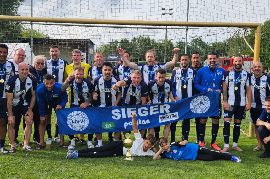 The photo shows a large group of people on a soccer field, positioned directly in front of a soccer goal. Most are wearing matching dark blue and white jerseys, dark shorts, and soccer cleats. Some people are standing in the back row, while others are in the front row, either crouching or kneeling. Several medals are visible.  A large blue banner is being held in the center of the group. It reads “SIEGER” in large white letters. On the left and right are association logos, including the logo of the Northeast German Soccer Association (NOFV) and the JFV. Other sponsor logos are also depicted on the banner.  In front of the banner, on the grass, stands a gold-colored trophy. Directly in front of it, two children in athletic wear are lying on their stomachs, propping up their upper bodies with their arms. Behind the group, the net of the soccer goal stretches across the goal. In the background, green spaces, trees, several buildings, and the technical facilities of a sports complex are visible. The sky is mostly cloudy. The photograph documents a sporting achievement and the joint celebration of a team following a competition.