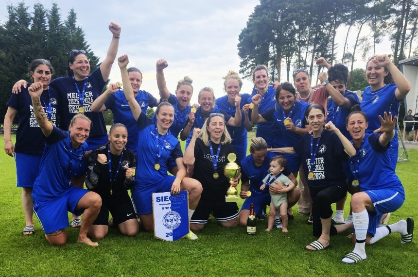 The photo shows a large group of people on an outdoor grass field. Most are wearing matching blue jerseys or dark blue shirts with the words “CHAMPIONS 2025/2026” and medals around their necks. The group is arranged in several rows; some people are standing, while others are kneeling on the grass. Many have one arm raised.  In the foreground, roughly in the center of the picture, stand a gold-colored trophy and a blue-and-white pennant. The word “SIEGER” and the logo of the Northeast German Soccer Association (NOFV) are clearly visible on the pennant. Next to the trophy, a bottle of sparkling wine sits on the grass.  To the right of the group is a toddler, who is also wearing a medal and is being supported by an adult. Most of the people are looking toward the camera.  The background consists of a well-maintained sports complex with green grass, tall trees, and a cloudy sky. The photo documents the award ceremony or celebration of a soccer victory.  Please provide your feedback on BizChat.