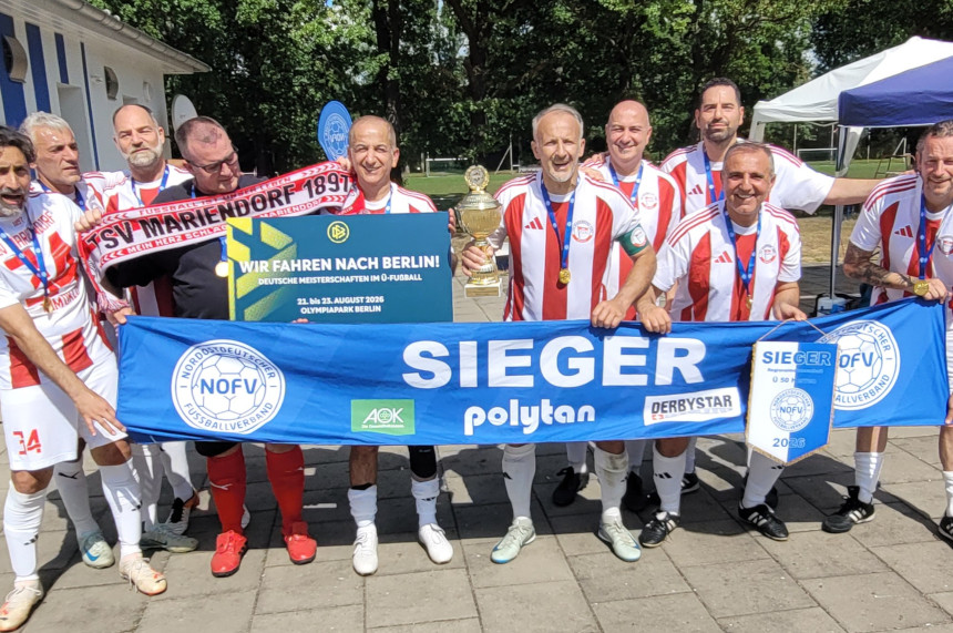 The photo shows a group of soccer players on a paved area outside a sports complex. Most of them are wearing matching white-and-red jerseys, white shorts, and medals around their necks. The players are standing close together and looking toward the camera.  In the foreground, the group is holding a large blue banner with the word “SIEGER” (WINNERS) on it. The banner also features the logos of the Northeast German Soccer Association (NOFV), AOK, Polytan, and Derbystar. To the right, there is also a blue winner’s pennant with the NOFV logo.  In the center of the image, a gold-colored trophy is on display. To the left of it, a person is holding a red-and-black fan scarf bearing the inscription “TSV Mariendorf 1897.” Next to it is a black sign that reads, among other things: “We’re heading to Berlin! German Over-50s Soccer Championships” and lists the dates “August 21–23, 2026, Olympiapark Berlin.”  The background shows trees, green spaces, a light-colored club building, and pavilions or tents set up. The photo documents the celebration of a successful over-50 soccer team and its qualification for the German Championships.