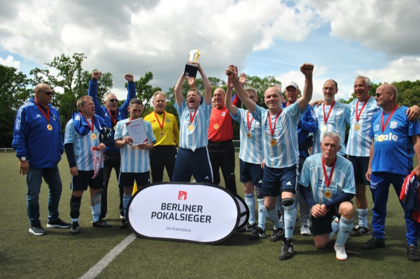 Das Bild wird analysiert: Gesichter sind möglicherweise verschwommen, um die Privatsphäre zu schützen. Eine Fußballmannschaft feiert mit Medaillen und Pokal. Ein Schild vor ihnen zeigt die Aufschrift „BERLINER POKALSIEGER“. Foto: BFV
