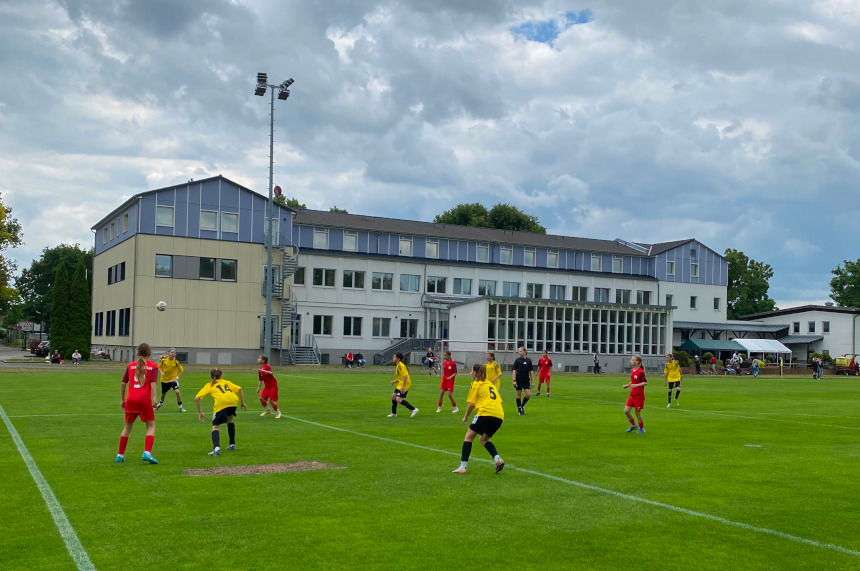Fußballspiel auf Rasenplatz vor großer Halle; Teams in Gelb und Rot, Ball im Spiel, mehrere Spielende verteilt, bewölkter Himmel. Foto: BFV