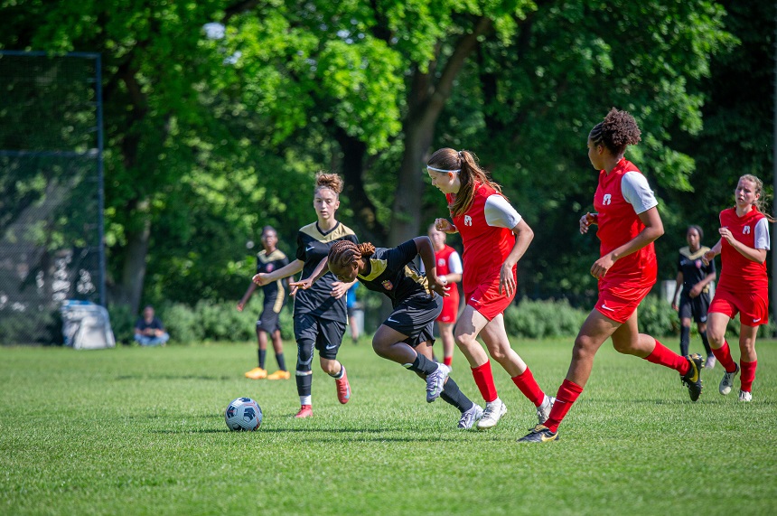 Das Foto zeigt eine dynamische Szene auf einem grünen Fußballfeld. Im Vordergrund sind vier junge Fußballspielerinnen zu sehen – zwei tragen rote Trikots, zwei schwarze. Eine Spielerin in schwarzem Trikot beugt sich nach vorne, um den Ball zu kontrollieren, während eine Spielerin in Rot versucht, den Ball zu erobern. Die Körperhaltungen deuten auf ein aktives Spielgeschehen hin. Im Hintergrund sind weitere Spielerinnen auf dem Feld verteilt sowie Bäume, die das Spielfeld umgeben.