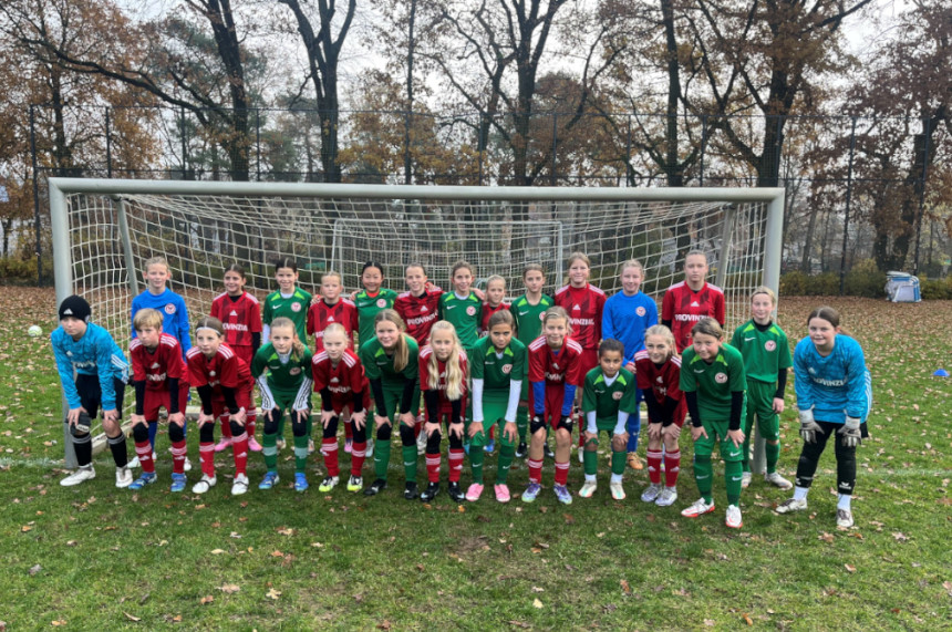 The picture shows a large group of children standing together on a soccer field. They are positioned in two rows in front of a soccer goal. The front row is kneeling slightly bent forward, while the back row is standing upright. The children are wearing different jerseys: some in red, others in green, and two in blue goalkeeper jerseys. All have soccer boots on, and some are wearing long socks in matching colors. The ground is covered with green grass, on which autumn leaves are scattered. Tall, bare trees can be seen in the background, hinting at an autumnal season. There is a wooded area behind the gate and the lighting appears natural, presumably with a cloudy sky.