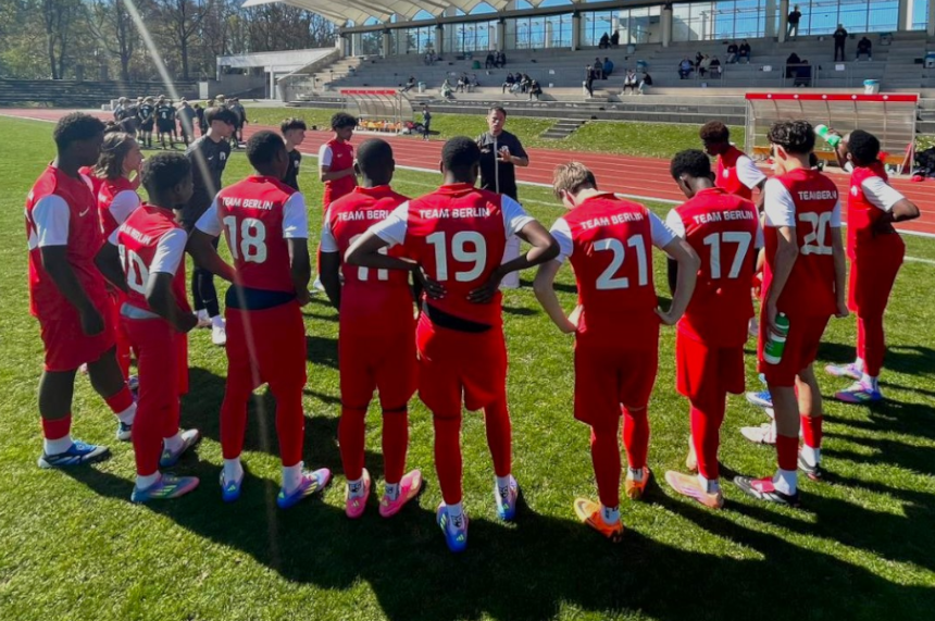 Das Foto zeigt eine Gruppe junger Fußballspielender, die auf einer grünen Rasenfläche im Freien im Kreis zusammenstehen. Alle tragen rote Sporttrikots und rote Hosen. Auf den Rückseiten der Trikots steht in weißen Buchstaben der Schriftzug „TEAM BERLIN“ sowie individuelle Rückennummern, zum Beispiel 18, 19, 21, 17 und 27. Viele der Spielenden tragen bunte Fußballschuhe und lange Sportsocken. Die Gruppe bildet einen engen Halbkreis um eine Person, die etwas erhöht vor ihnen steht und spricht. Diese Person trägt dunkle Kleidung und hält die Hände vor dem Körper. Die Spielenden stehen mit dem Rücken zur Kamera, einige haben die Hände in die Hüften gestützt, andere halten Trinkflaschen oder haben die Arme locker hängen. Im Hintergrund ist ein Sportstadion zu sehen. Dort gibt es eine Laufbahn, eine überdachte Tribüne mit Sitzplätzen sowie einige Menschen, die verteilt auf den Rängen sitzen. Rechts am Bildrand steht eine rote überdachte Ersatzbank. Hinter dem Stadion sind Bäume zu erkennen, die Sonnenlicht fällt schräg von links ins Bild und wirft lange Schatten auf den Rasen.