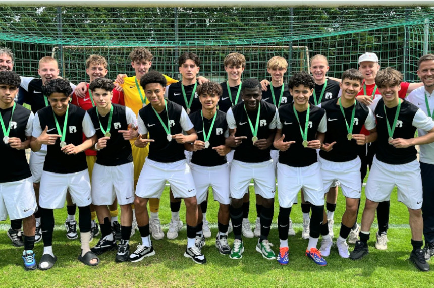 Das Foto zeigt eine Gruppe von 16 jungen Fußballspielern, die gemeinsam vor einem Tor auf einem grünen Rasenplatz stehen. Alle tragen schwarz-weiße Trikots und haben silberne Medaillen um den Hals – ein Hinweis auf eine erfolgreiche Teilnahme an einem Turnier oder Spiel. Einige der Spielerinnen haben die Arme freundschaftlich umeinander gelegt. Im Hintergrund ist das Tornetz sowie grüne Vegetation zu sehen. Die Gruppe wirkt geschlossen und stolz, was auf ein Gemeinschaftsgefühl und Teamgeist hindeutet.
