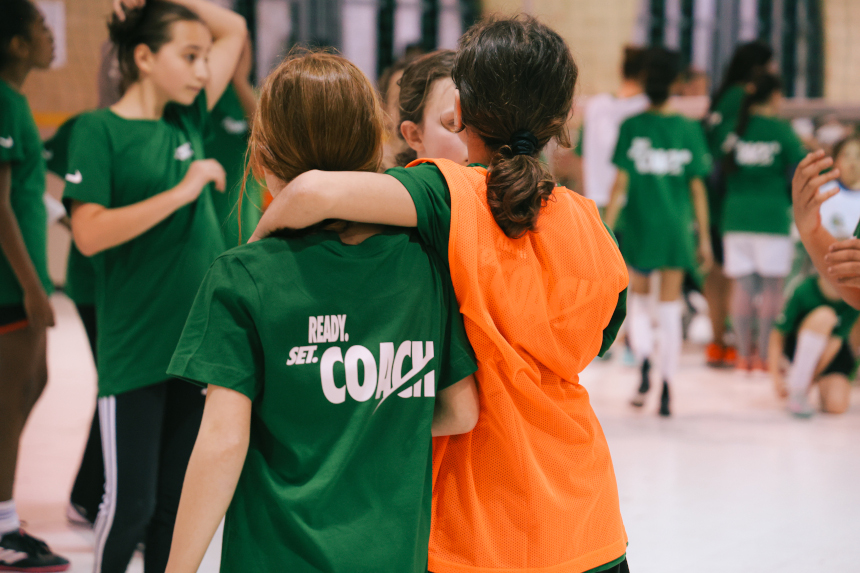 Kinder trainieren in einer Halle; zwei Kinder stehen im Vordergrund eng nebeneinander, umgeben von weiteren spielenden Kindern in grünen Shirts. Foto: Lea Gleisberg