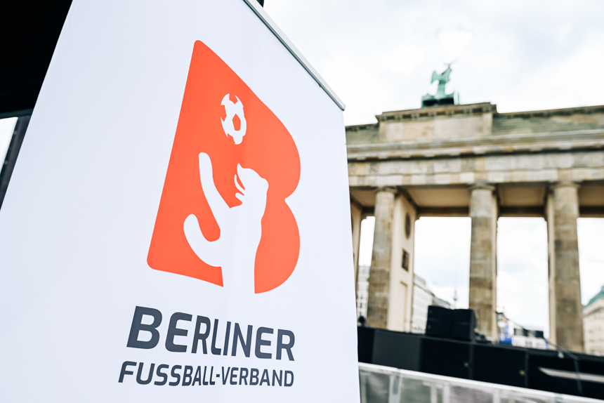 Im Vordergrund steht eine weiße Infotafel mit dem Logo des Berliner Fußball-Verbandes. Das Logo zeigt einen stilisierten roten Buchstaben „B“, in den die Silhouette eines aufrecht stehenden Bären integriert ist. Über der Pfote des Bären schwebt ein Fußball. Unter dem Logo steht der Schriftzug „Berliner Fußball-Verband“. Im Hintergrund ist unscharf das Brandenburger Tor zu sehen. Die hellen Sandstein-Säulen des historischen Bauwerks sind gut erkennbar, und oben auf dem Tor befindet sich die Quadriga, eine vierspännige Wagenfigur. Der Himmel ist leicht bewölkt. Die Gesamtstimmung wirkt hell und klar.