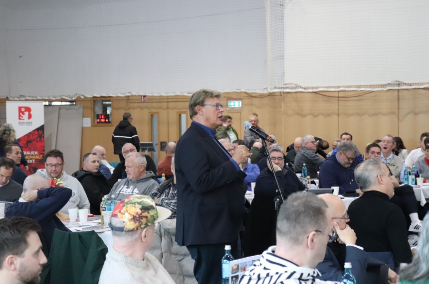 Person stands in a hall between round tables and speaks into a microphone, surrounded by many seated participants. Photo: BFV