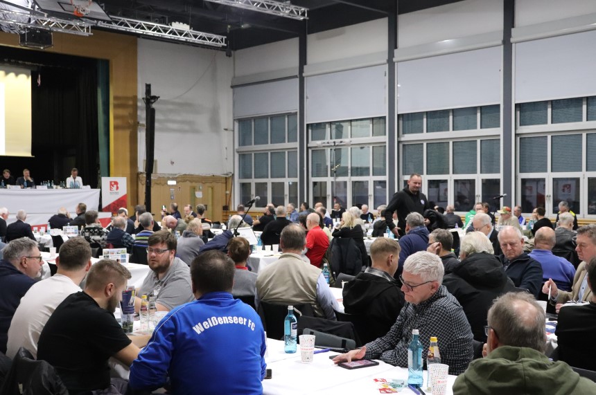 Large hall with many people at round tables, podium with speakers in the front, large windows and bright lighting in the background. Photo: BFV
