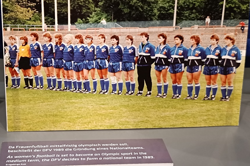 Das Foto zeigt eine Frauenfußballmannschaft, die nebeneinander auf einem Fußballfeld steht. Die Spielerinnen tragen blaue Trikots mit weißen Streifen sowie blaue Shorts. Eine Spielerin hebt sich durch ein gelbes Trikot hervor – sie ist vermutlich die Torhüterin. Im Hintergrund sind Tribünen und grüne Bäume zu erkennen, was auf ein Stadion oder Sportplatz im Freien hinweist.  Im oberen Bereich des Bildes befindet sich ein Text in Deutsch und Englisch:  Deutsch: „Da Frauenfußball mittelfristig olympisch werden soll, beschließt der DFV 1989 die Gründung eines Nationalteams.“ Englisch: „As women's football is set to become an Olympic sport in the medium term, the DFV decides to form a national team in 1989.“