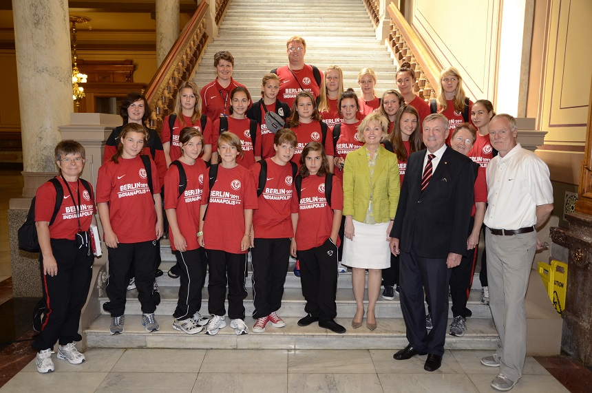 Das Foto zeigt eine Gruppe von etwa 20 Menschen, die auf einer breiten Marmortreppe stehen und in die Kamera lächeln. Die meisten Personen sind Jugendliche und tragen rote T-Shirts. Sie haben überwiegend schwarze Hosen und Sportschuhe an. In der Mitte der Gruppe stehen zwei Erwachsene: eine Frau mit einem gelben Blazer und weißem Rock sowie ein Mann im dunklen Anzug mit roter Krawatte. Rechts außen steht ein weiterer Erwachsener in einem weißen Polohemd und beiger Hose. Die Gruppe wirkt fröhlich und stolz.